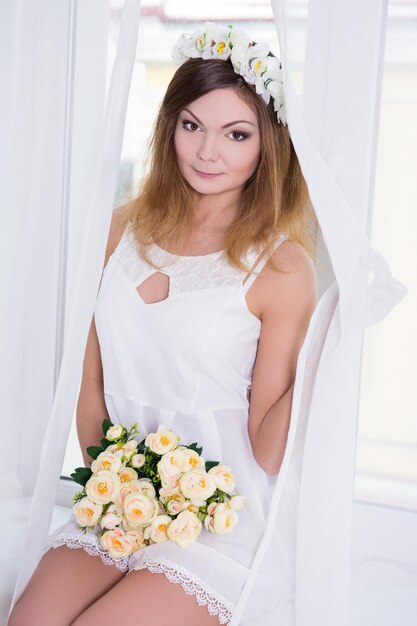 Portrait of young beautiful woman in short white bridal dress with flowers sitting on window sill