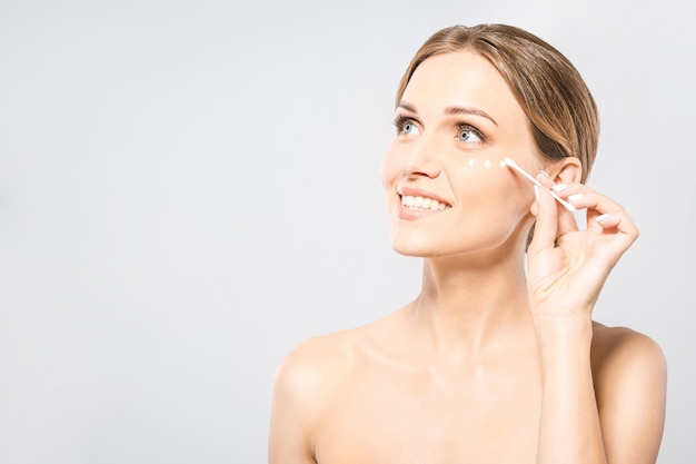 Portrait of young beautiful woman smiling while taking some facial cream using a cotton swab isolated on white background with copy space.