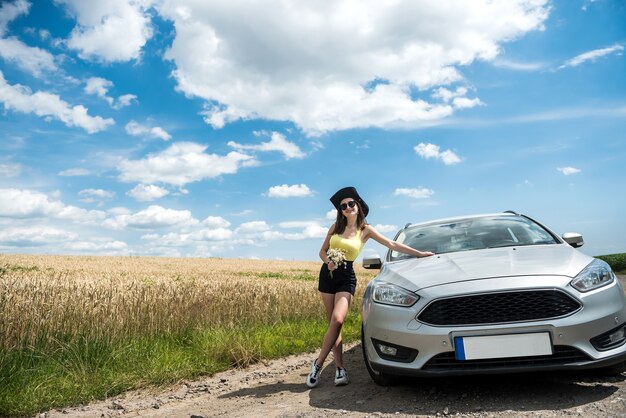 Portrait of young beautiful woman standing near her car at rural road. Dream for perfect trip in summer time