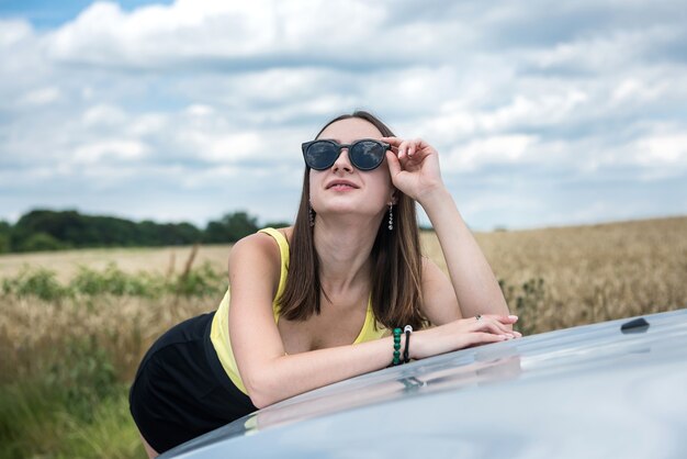 田舎道で彼女の車の近くに立っている若い美しい女性の肖像画。夏の完璧な旅行を夢見てください