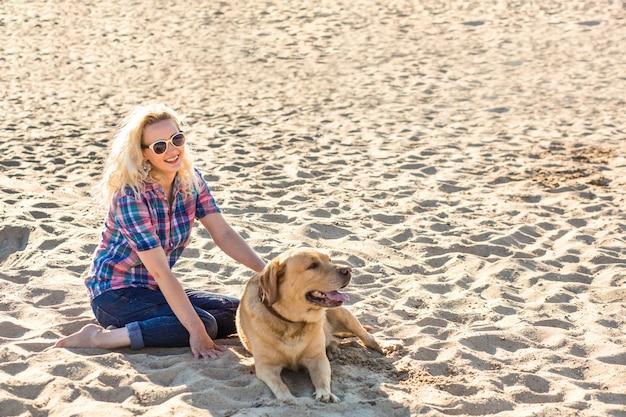 Portrait of young beautiful woman in sunglasses sitting on sand beach with golden retriever dog girl...