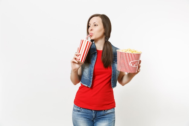 Portrait of young beautiful woman watching movie film, holding bucket of popcorn, drinking from plastic cup of soda or cola looking aside on copy space isolated on white background. Emotions in cinema