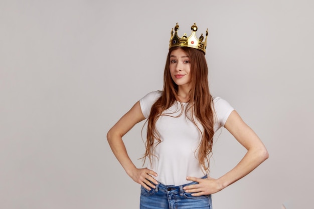 Photo portrait of young beautiful woman with crown on head keeping hands on hips, self-motivation and dreams to be best, wearing white t-shirt. indoor studio shot isolated on gray background.