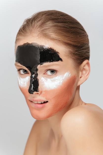 Portrait of young beautiful woman with different cosmetic masks on face biting lip while dreamily looking in camera over gray background