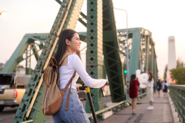 近代的な都市の背景に橋を渡る電動スクーターを持つ若い美しい女性の肖像画