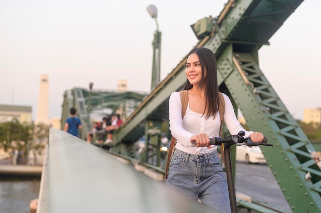 近代的な都市の背景に橋を渡る電動スクーターを持つ若い美しい女性の肖像画