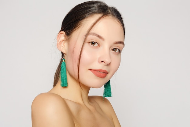 Portrait of young beautiful woman with perfect skin and bright make-up touching her face with manicured fingers. Long green fabric earrings in her ears. Her shoulders naked. Close up. Copy-space