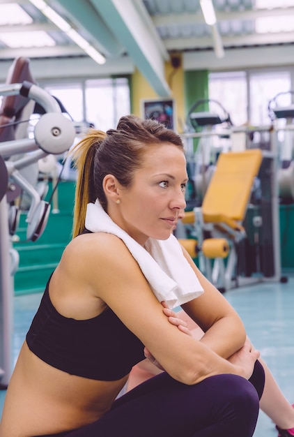 Ritratto di giovane bella donna con asciugamano che riposa seduta sul pavimento del centro fitness dopo una dura giornata di allenamento