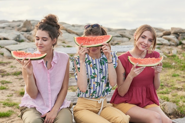 Portrait of young beautiful women holding slices of watermelon and smiling at camera cheerfully while spending summer evening outdoors