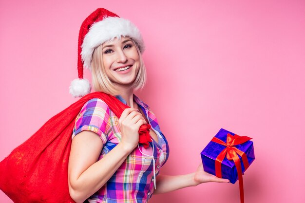 Portrait of young Beautyful blond amazed woman with christmas box gift on red background. lady in a plaid shirt and santa claus hold a bag full of presents on a pink background in the studio.