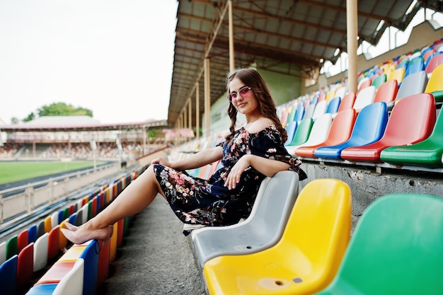 Portrait of a young beutiful girl in dress and sunglasses sitting on the tribunes in stadium