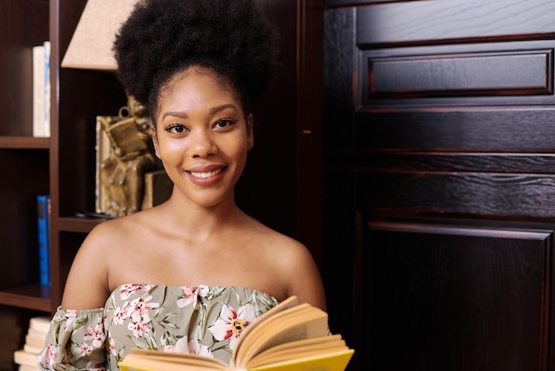 Portrait of a young black female student. She is standing in the university library, holding a book and looking at the camera.