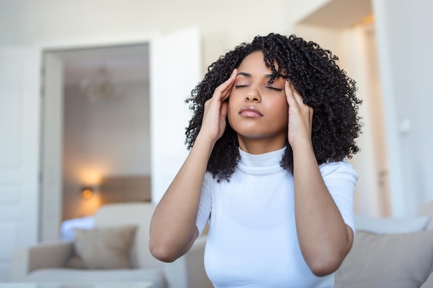 Portrait of a young black girl sitting on the couch at home\
with a headache and pain beautiful woman suffering from chronic\
daily headaches sad woman holding her head because sinus pain