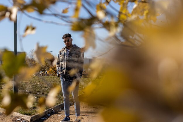 Photo portrait of a young black latino man with sunglasses walking along a road in summer view from behind the leaves of a tree