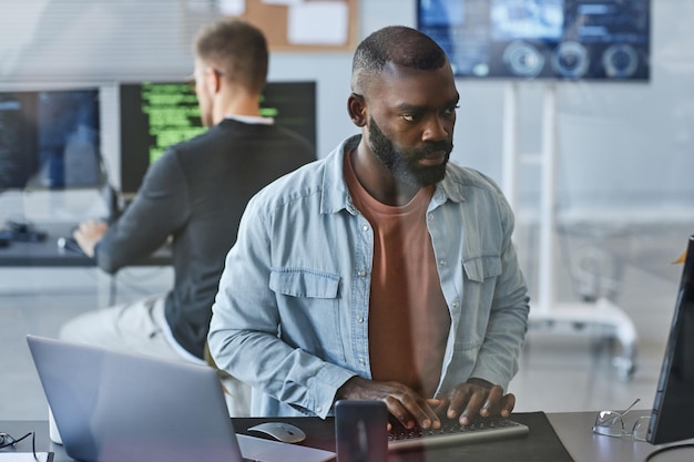 Portrait of young black man using computer while programming software in it company office behind gl