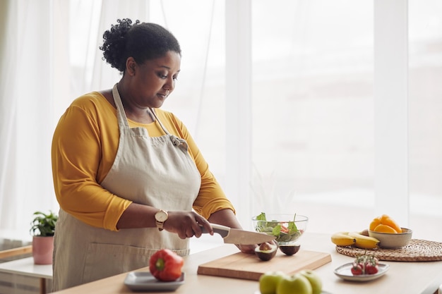 Portrait of young black woman cutting avocado while making salad in sunlit kitchen copy space