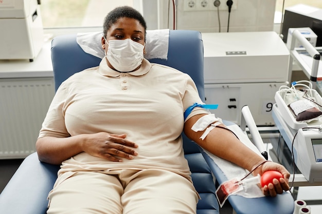 portrait of young black woman donating blood while lying in chair and wearing mask copy space