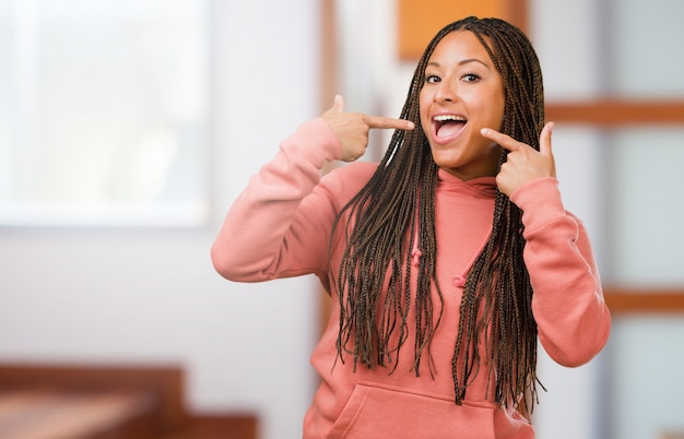 Portrait of a young black woman wearing braids smiles, pointing mouth, concept of perfect teeth, white teeth, has a cheerful and jovial attitude