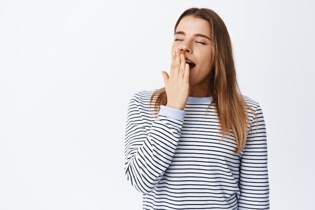 Portrait of young blond girl waking up sleepy close eyes and yawning with opened mouth cover it with hand standing tired against white background