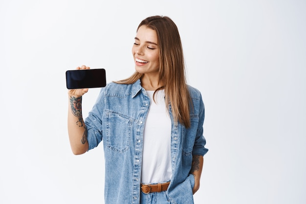 Портрет молодой белокурой женщины, держащей телефон горизонтально, показывая пустой экран смартфона для рекламы приложения, стоящей над белой стеной