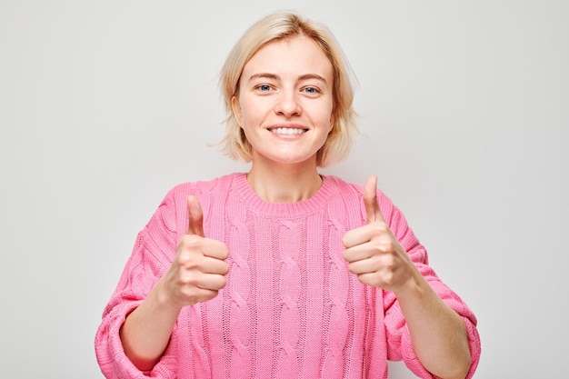 Photo portrait of young blond woman in pink sweater smiling joyfully showing thumbs up gesture isolated on white studio background approves good choice right decision