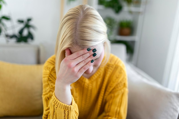 頭痛と片頭痛で自宅のソファに座っている若いブロンドの女性の肖像画。慢性的な毎日の頭痛に苦しんでいる美しい女性。副鼻腔の痛みのために頭を抱えている悲しい女性