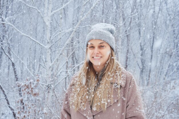 降雪時の森の冬のコートと帽子の若いブロンドの肖像画。