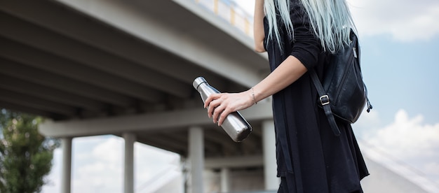 Portrait of young blonde girl, dressed in black with backpack, holding steel thermo bottle, under the modern bridge.