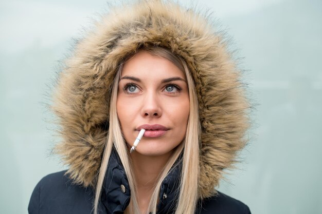 흰색 배경 위에 격리된 입에 후드와 담배를 넣은 겨울 재킷을 입은 젊은 금발 소녀의 초상화