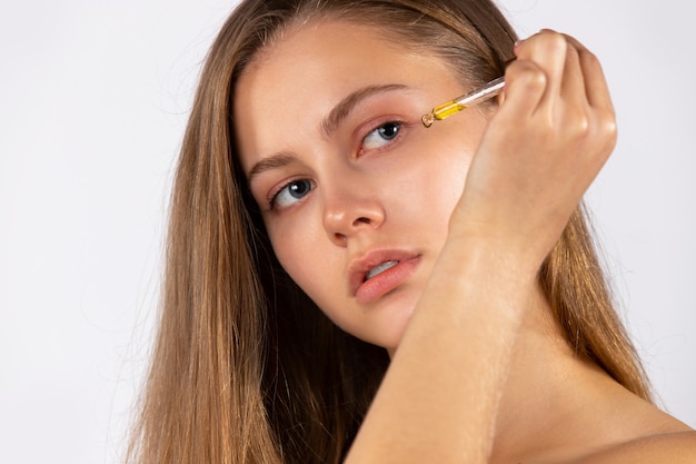 Portrait of a young blonde girl without makeup applies a serum to her face with a pipette. Cosmetology, skin care, spa, natural cosmetics. Photo on white wall. High quality photo