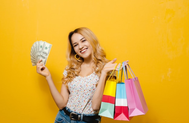 Ritratto di una giovane ragazza bionda felice in possesso di banconote e shopping bag sul muro giallo