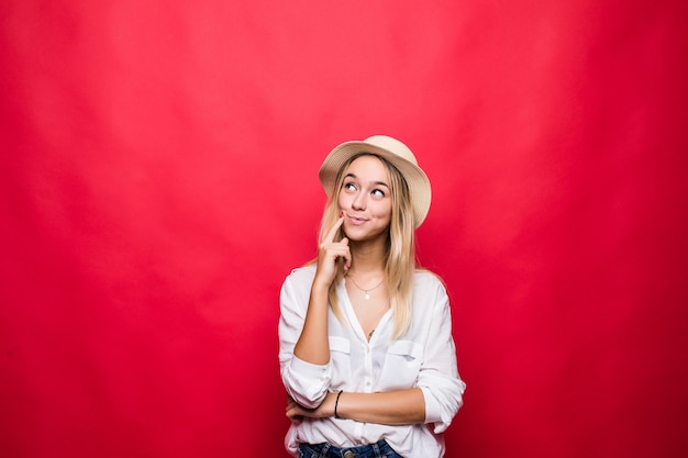 Portrait of young blonde woman wearing in straw hat and touching chin with brooding or dreaming view, isolated over red wall