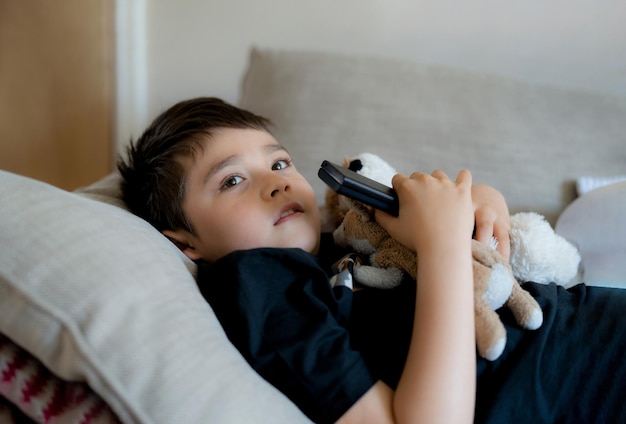 Portrait young boy holding remote control and looking up with curious face Kid sitting on sofa watching cartoon on TV Child lying down on couch relaxing in living room after back from school