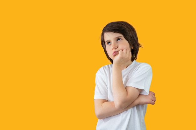 Portrait of a young boy of nine years old wearing white t-shirt, pressing his hand to his face, thinking seriously and looking up