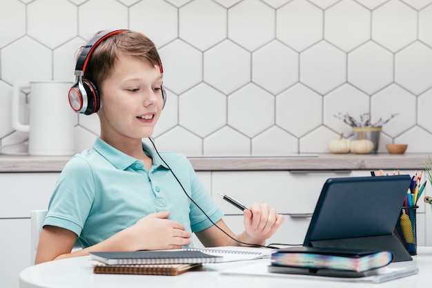 Portrait of young boy sitting at desk near notebooks looking at screen of tablet having online lesson holding pen