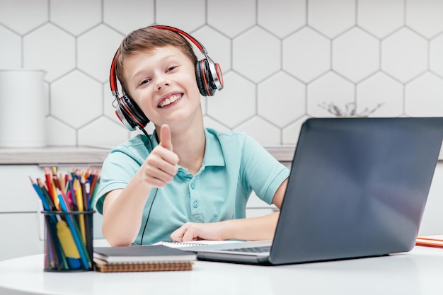 Portrait of young boy wearing black red headphones sitting at\
desk in front of laptop showing thumb up sign gesturing