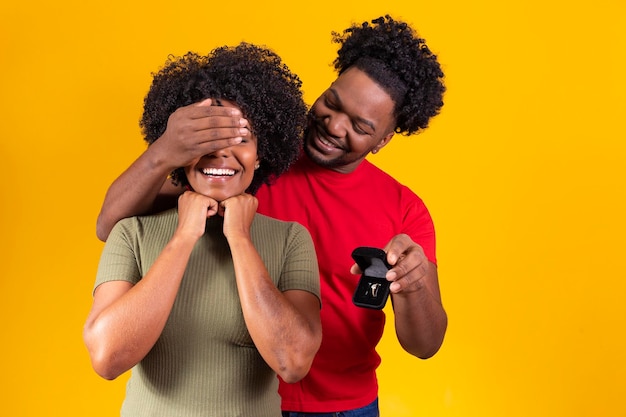 Portrait of young boyfriend holding small box with jewelry\
isolated yellow background covering his girlfriend\'s eyes