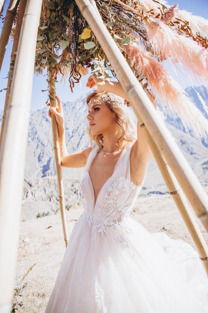 乾燥した花の結婚式のアーチの反対側の写真のためにポーズをとっているスタイリッシュなウェディングドレスに身を包んだ若い花嫁の肖像画セクシーで神秘的な外観完璧なメイク