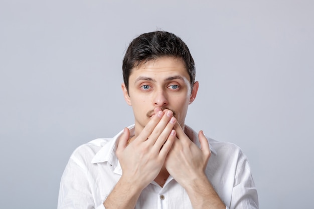 Portrait of a young Brunet man in white shirt covers his mouth with his hands on gray background. guy knows the secret but won't tell