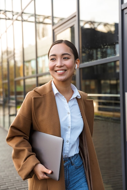 Portrait of young brunette asian woman wearing earbuds holding laptop computer while walking through city street