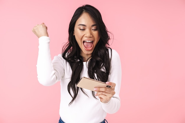 Portrait of young brunette asian woman with long hair holding cellphone and playing video games isolated over pink wall