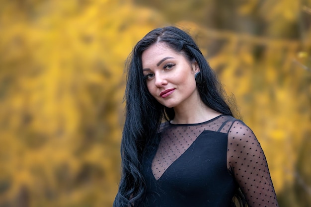 Portrait of a young brunette girl with long black hair and in a black dress in the autumn park