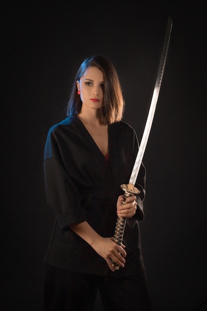 Portrait of a young brunette kung fu girl with a Japanese sword on a black background