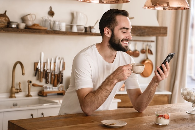 自宅のスタイリッシュなキッチンで朝食をしながらコーヒーを飲み、スマートフォンを使用して若いブルネットの男の肖像画