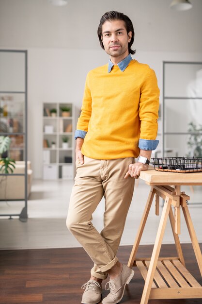 Portrait of young brunette man in stylish outfit standing at wooden table in modern home office