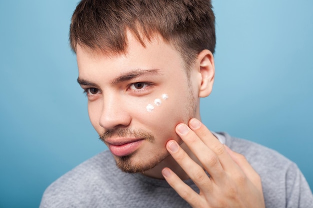 目の治療または保湿化粧品のスキンケアの概念の下で顔のクリームを適用する小さなひげと口ひげを持つ若いブルネットの男の肖像画青い背景で隔離の屋内スタジオショット