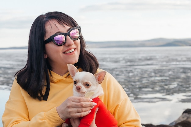 Portrait of a young brunette in nature with a Chihuahua dog in her arms. The girl smiles at the camera.