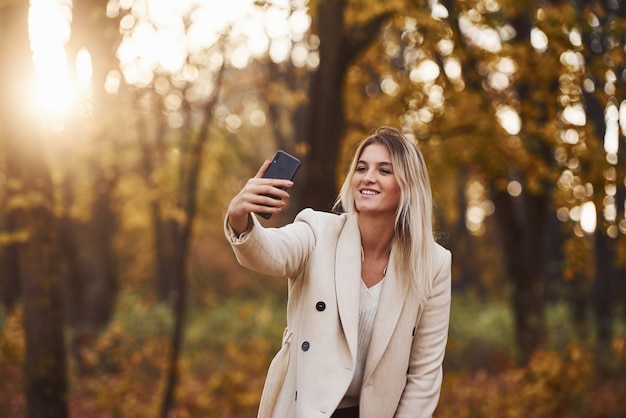 Portrait of young brunette with phone in hand that is in autumn forest at daytime.