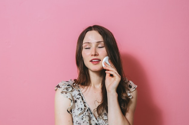 Portrait of young brunette woman cleaning her face with a cotton pad