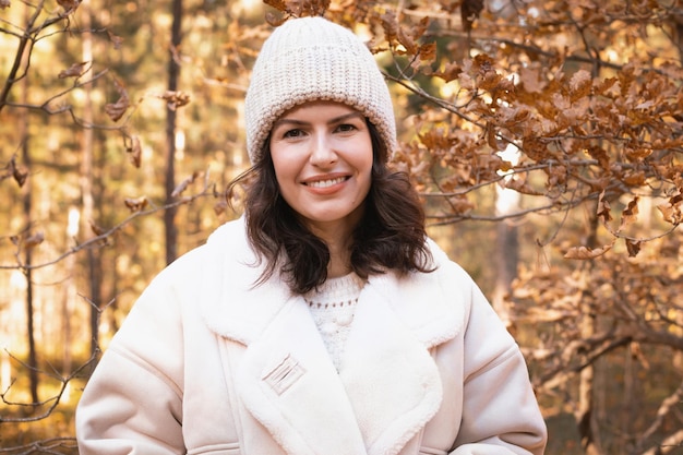 Portrait of a young brunette woman in a knitted hat and white jacket in the autumn forest